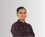 Seorang pria berwajah buram berdiri dengan lengan disilangkan, mengenakan kemeja kotak-kotak merah dan biru, di depan latar belakang gradien abu-abu ke putih.