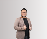 Seorang pria dewasa berjanggut mengenakan blazer cokelat dan kaos hitam, tersenyum sambil merapikan blazer, berdiri di depan latar belakang polos bergradasi abu-abu.