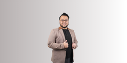 Seorang pria dewasa berjanggut mengenakan blazer cokelat dan kaos hitam, tersenyum sambil merapikan blazer, berdiri di depan latar belakang polos bergradasi abu-abu.